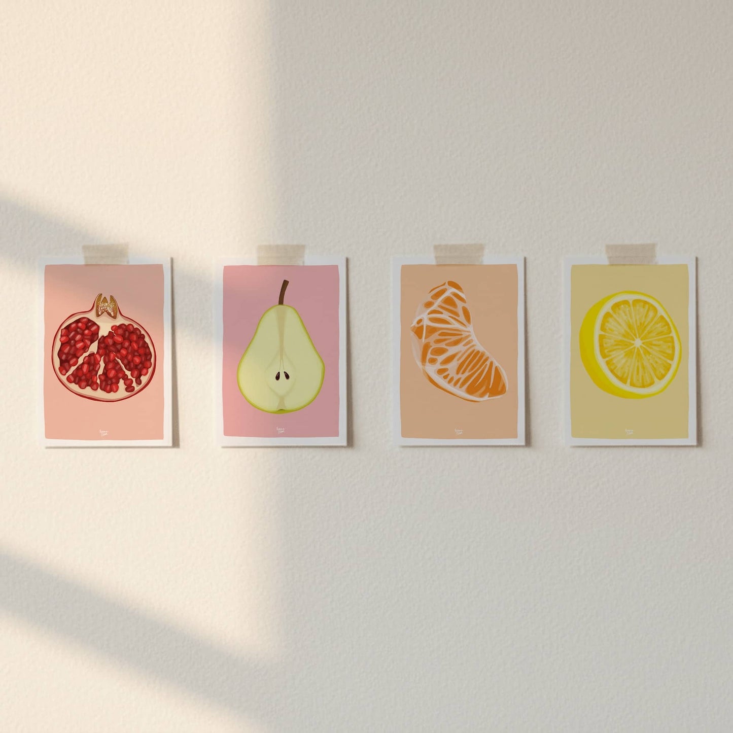 Cuatro postales de frutas pegadas con cinta en una pared blanca con luz natural lateral.