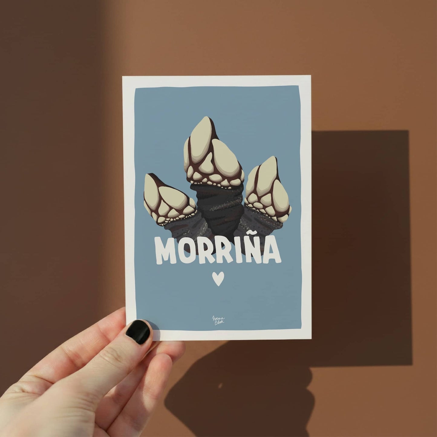 Una mano sostiene la mini lámina morriña para mostrar el tamaño A6 y el marco blanco que ayuda al proceso de enmarcación por normablok.