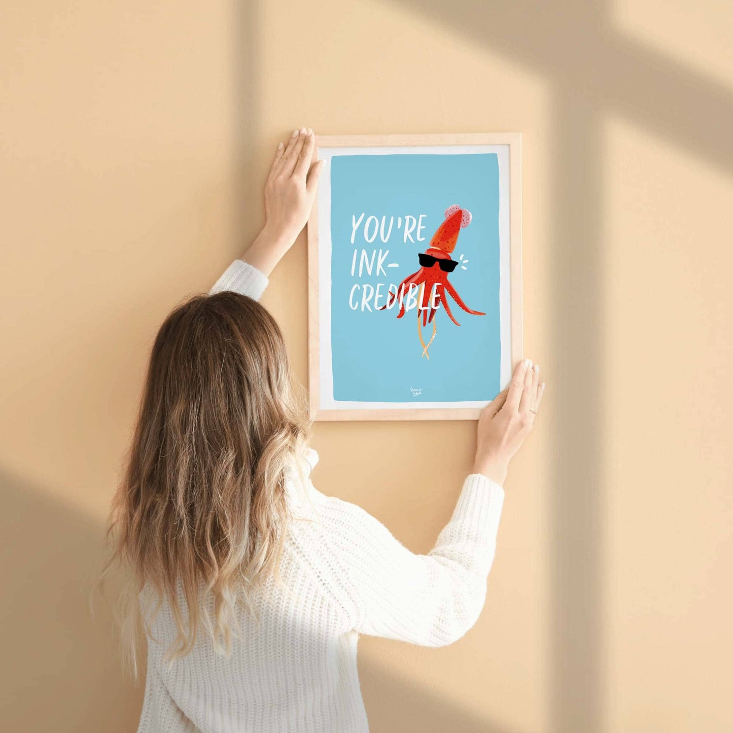 A woman hangs the A4-sized framed squid art print on a neutral wall. Her hands are adjusting the frame, providing a real-life reference for the physical scale and proportions of the printed illustration, making it easy to visualize the size for a bedroom or studio wall.