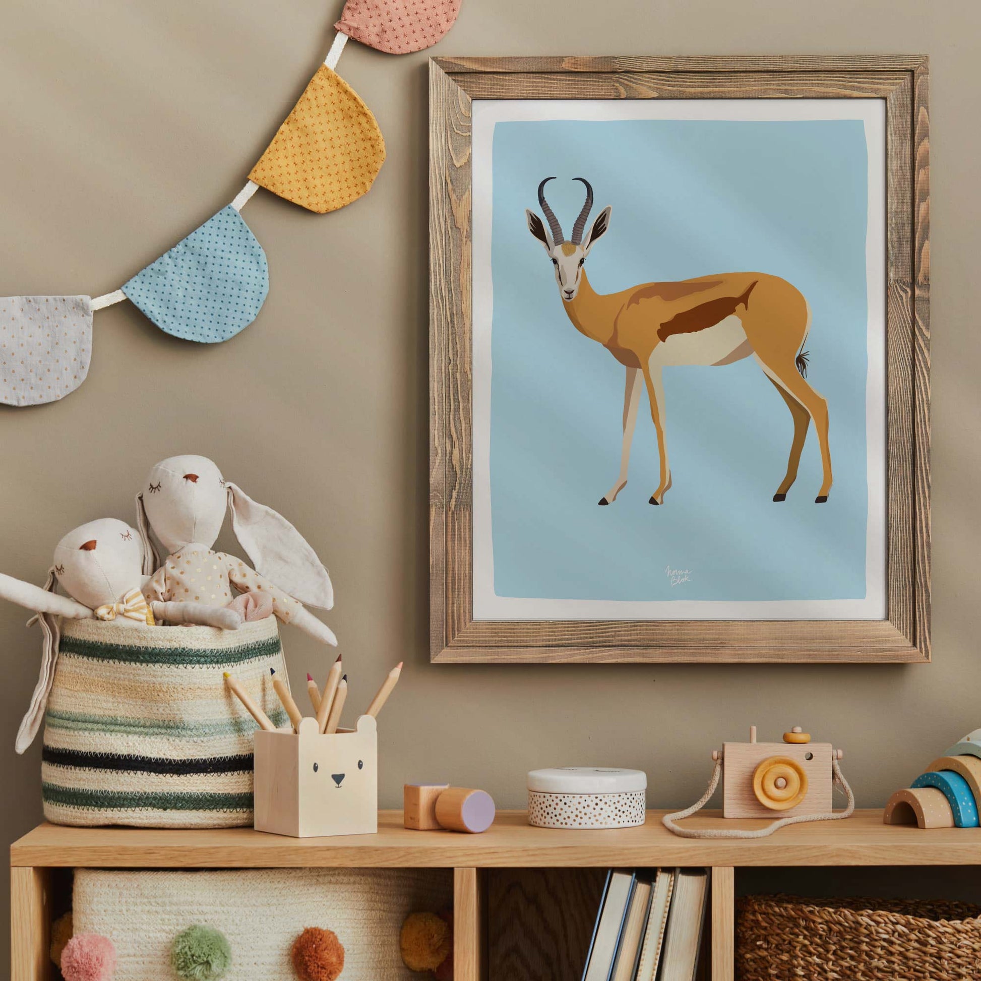 The antelope art print displayed in a charming vintage frame within a cozy nursery. Placed on a wooden shelf surrounded by toys and plushies, this setup demonstrates how wildlife illustrations create a warm, inviting, and playful environment for children.