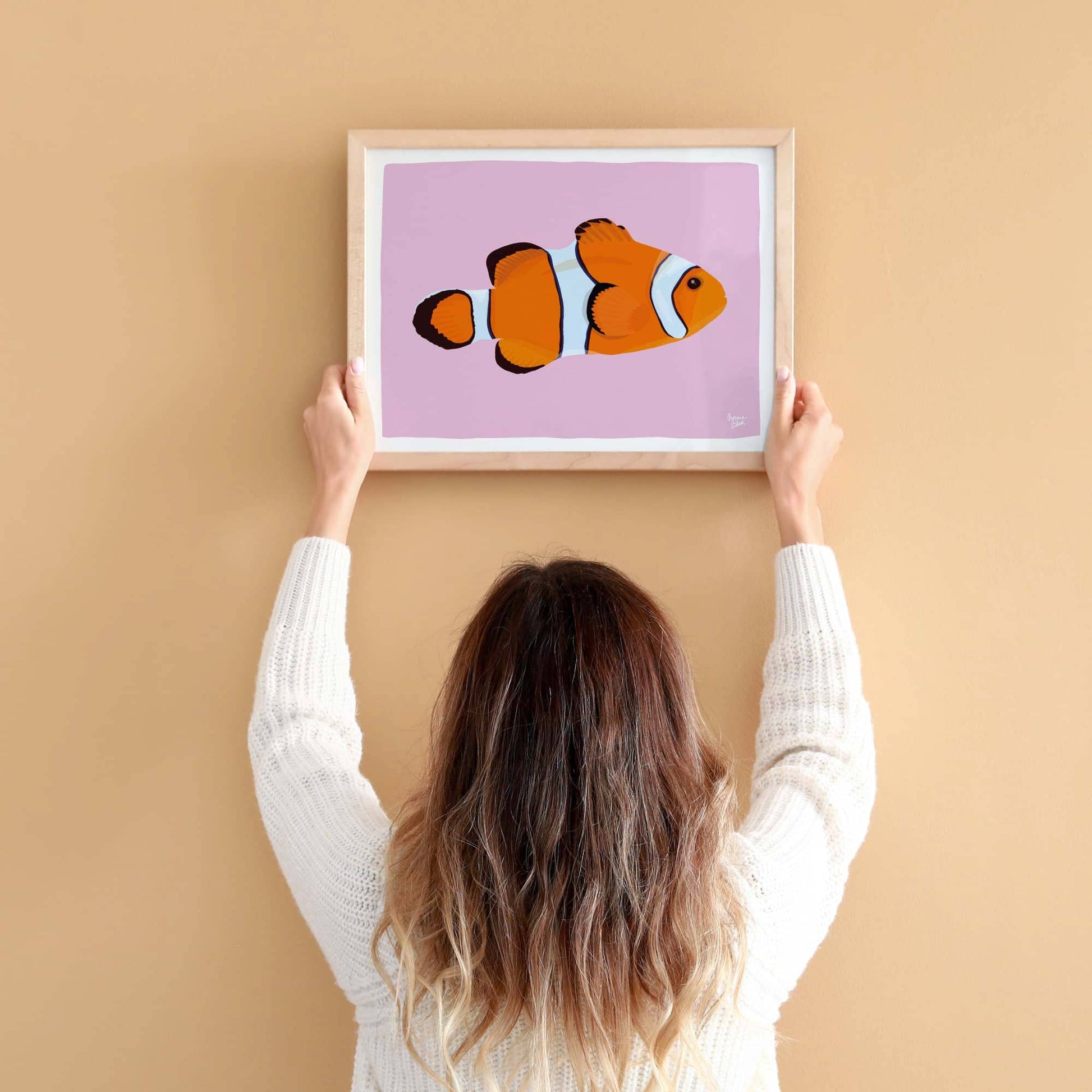A woman hangs an A4-sized picture of a clownfish on a light orange wall. It helps to see the true scale of the illustration and how the lilac background stands out against the warm wall.