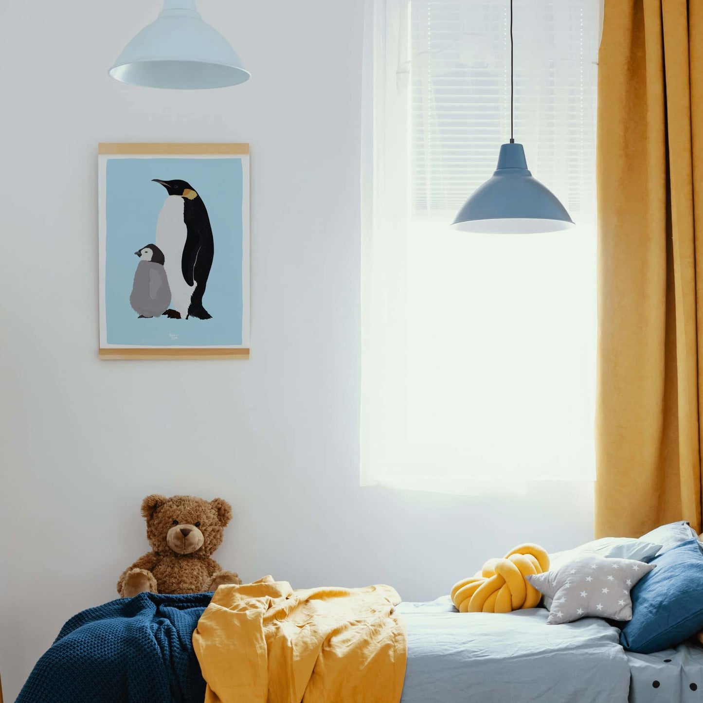 Emperor penguin print hanging with a magnetic wooden frame in a kids' room with blue and yellow bedding and a plush bear toy.