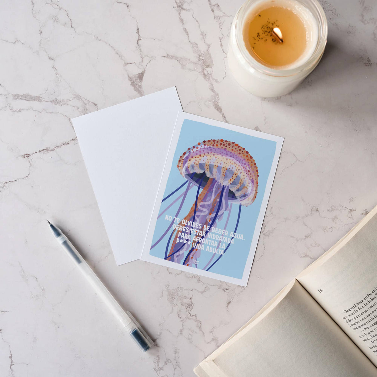 Vista cenital de una mesa acogedora con un libro, una vela y dos postales de la medusa, mostrando el diseño frontal y la parte trasera totalmente en blanco.