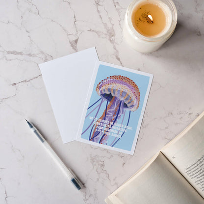 Vista cenital de una mesa acogedora con un libro, una vela y dos postales de la medusa, mostrando el diseño frontal y la parte trasera totalmente en blanco.