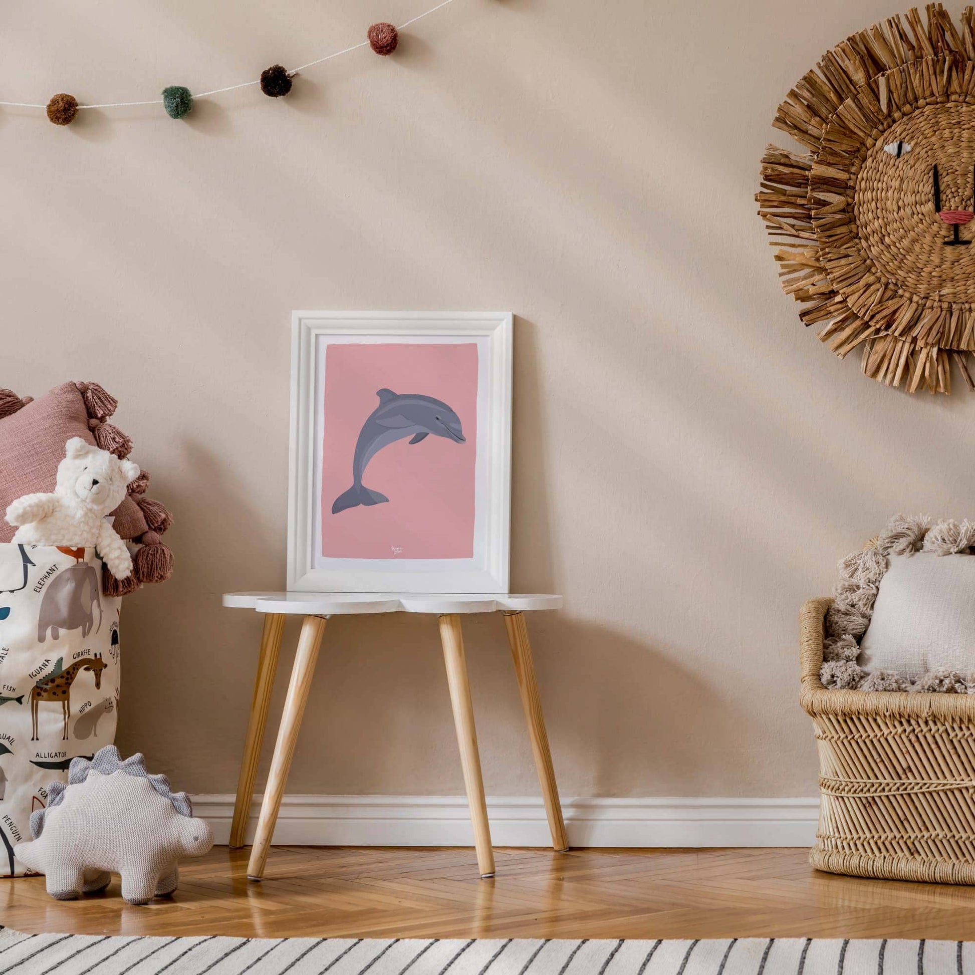 Lámina del delfín en un marco blanco minimalista colocada encima de una mesita en una habitación infantil llena de peluches de animales. Los juguetes que la rodean enfatizan la naturaleza juguetona y temática de la decoración de la habitación.