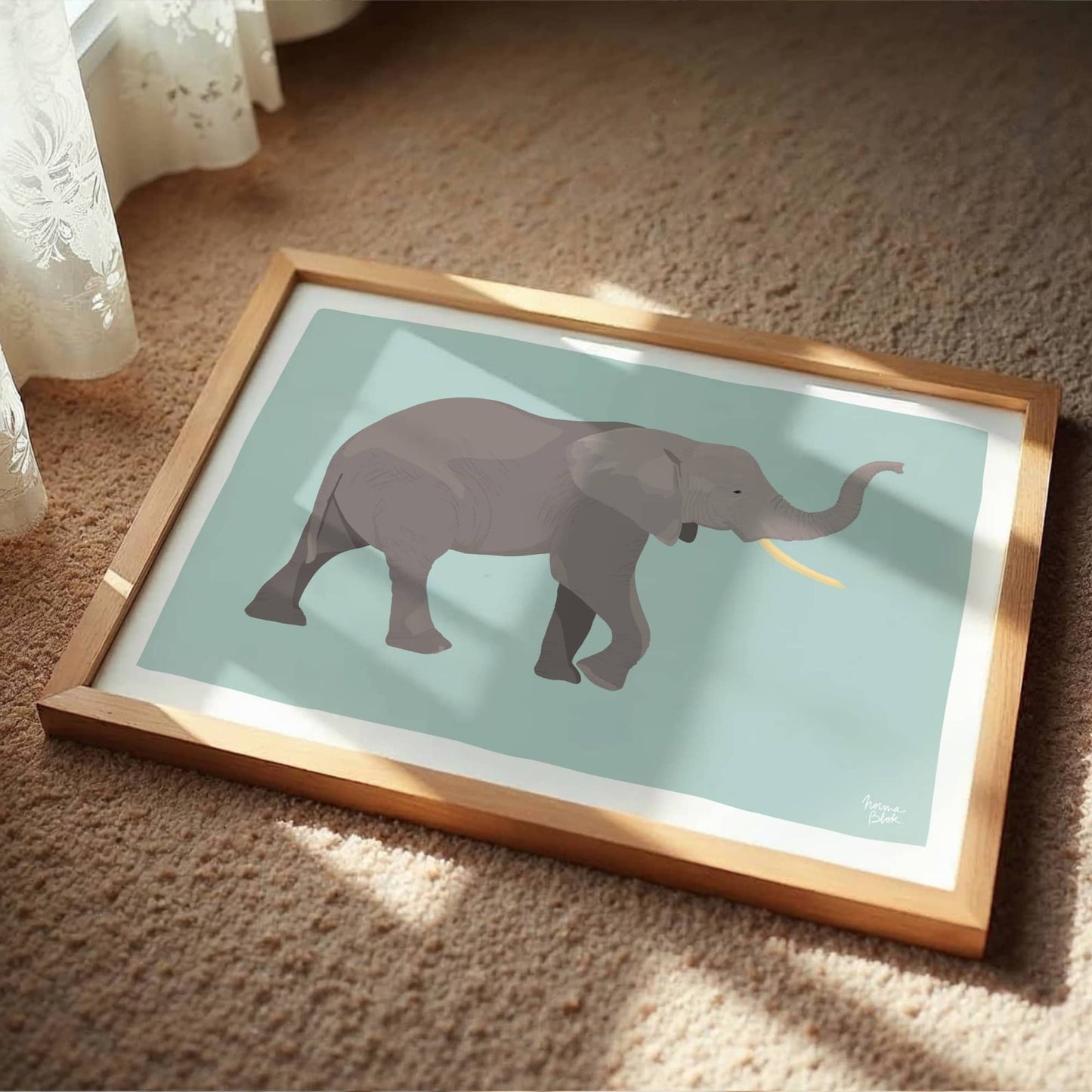 The African elephant art print in a natural wooden frame sitting casually on a soft carpet floor next to a flowing window curtain. This cozy, minimalist setting provides a modern styling idea for a warm nursery, kids' playroom, or relaxed bedroom corner.