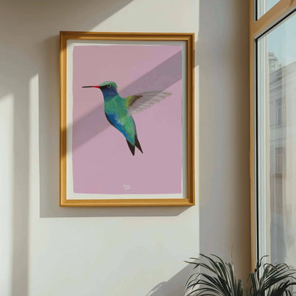 La lámina del colibrí en un marco minimalista colocada junto a una ventana luminosa por donde entra luz natural. La luz del sol acentúa los detalles vibrantes turquesa y rosa de la ilustración, mostrando la alta calidad de los colores y la nitidez de las líneas.