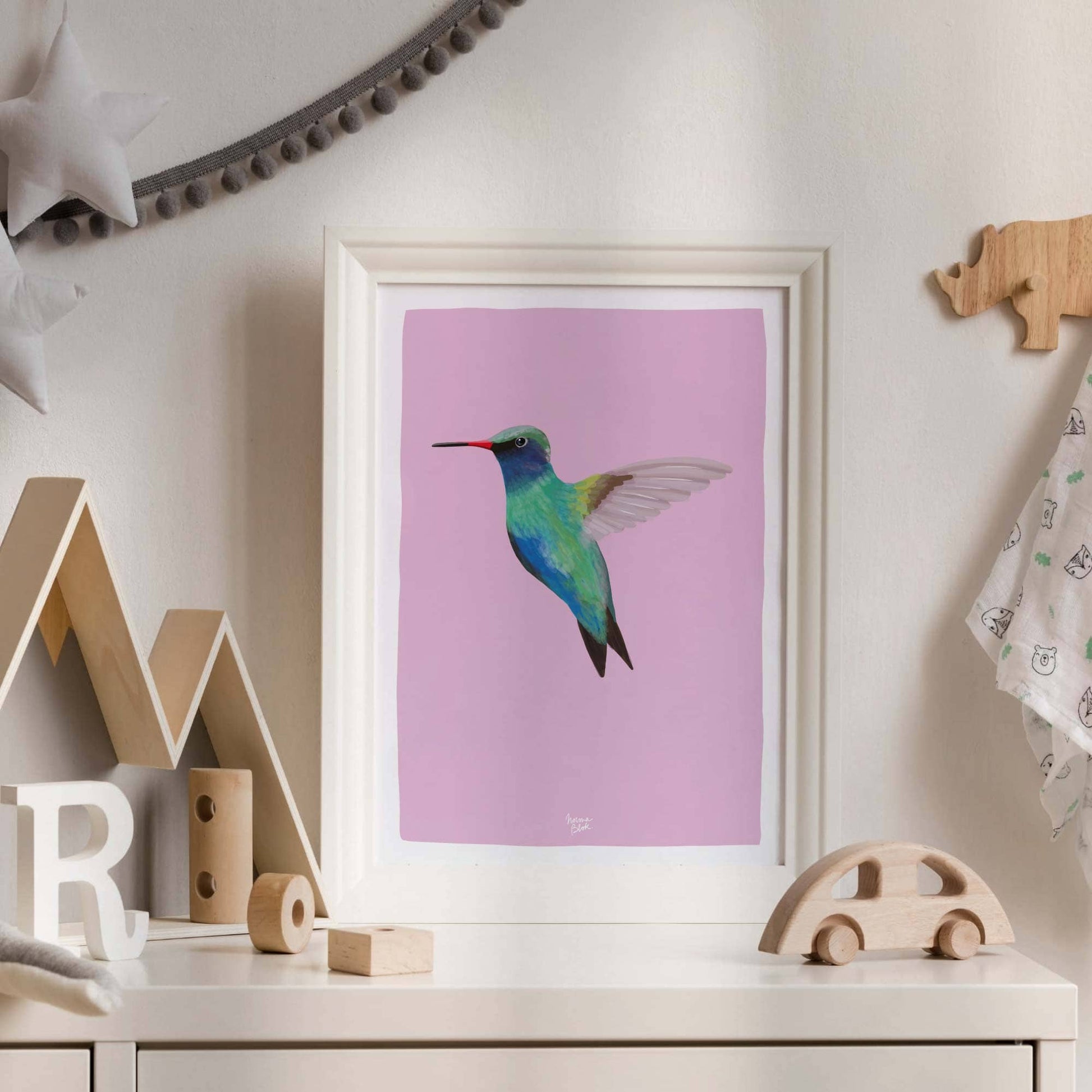 The hummingbird print in an elegant white molding frame, leaning against the wall on a white furniture piece in a nursery. The clean, bright white furniture makes the violet, turquoise, and pink colors of the bird stand out beautifully for a fresh look.