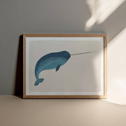 A framed narwhal illustration resting on a sand-colored floor against a warm, light grey-beige wall, highlighting a natural and earthy decor style.