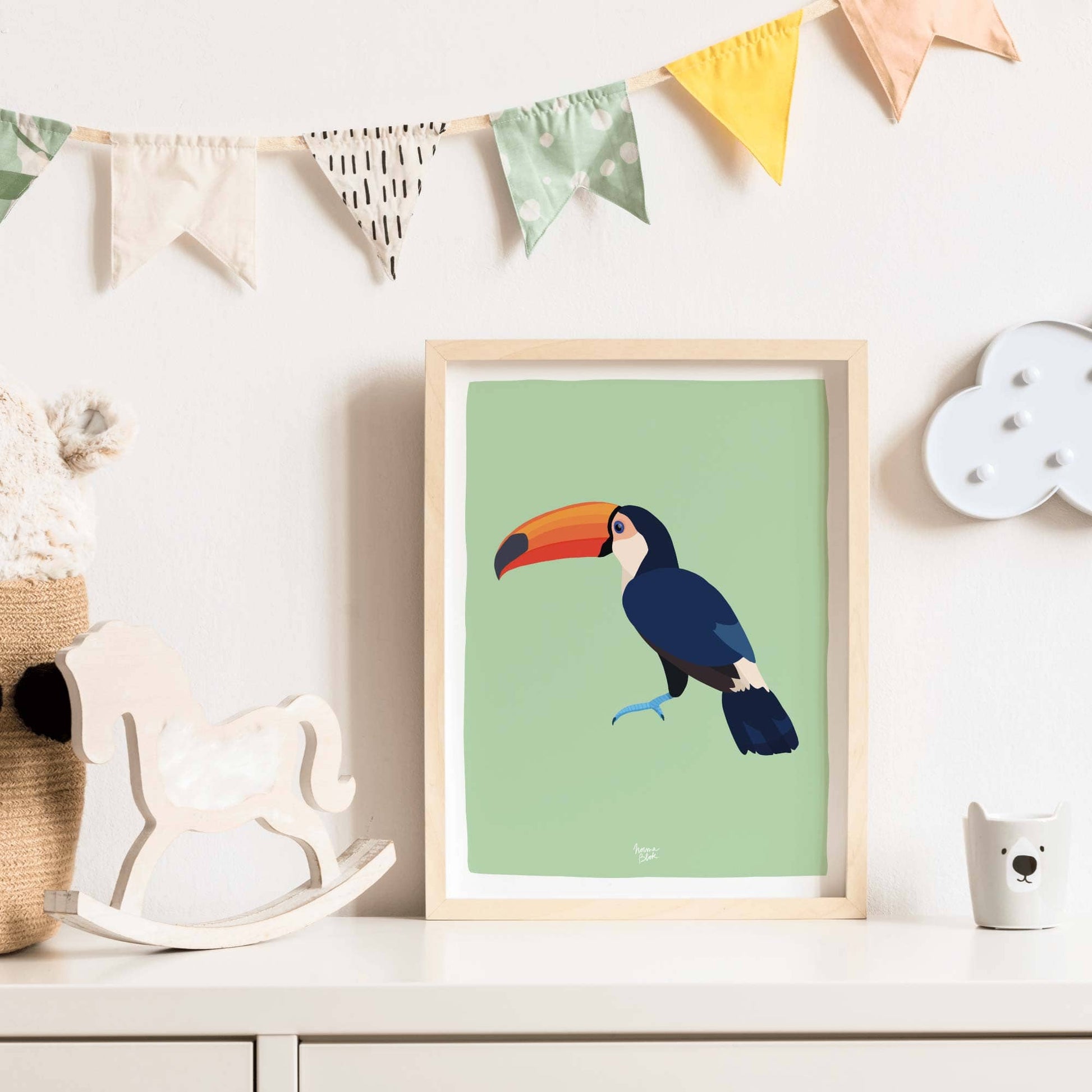 The toucan art print in a light wood frame leaning against the wall on a white dresser in a kids' room. The combination of light wood and bright white furniture creates a clean, fresh, and modern nursery aesthetic that highlights the toucan's vibrant colors.