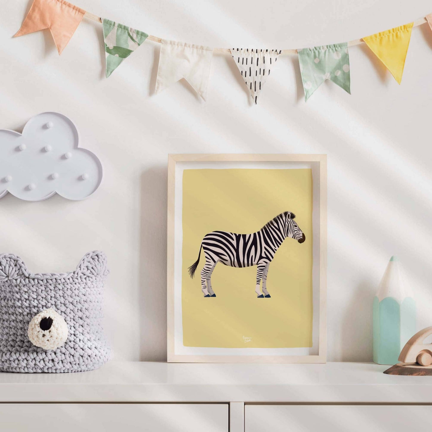 The framed zebra art print hanging in a bright white children's bedroom decorated with colorful fabric bunting. The yellow background of the zebra print adds a warm, playful touch that perfectly complements the cheerful and festive nursery atmosphere.