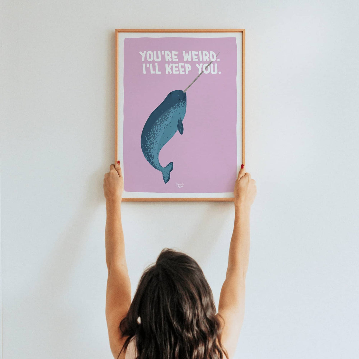 A lifestyle shot of a woman holding up a framed A3 size narwhal art print in a real room by Normablok.