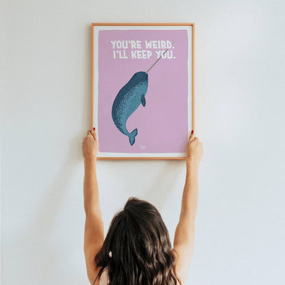 A lifestyle shot of a woman holding up a framed A3 size narwhal art print in a real room by Normablok.