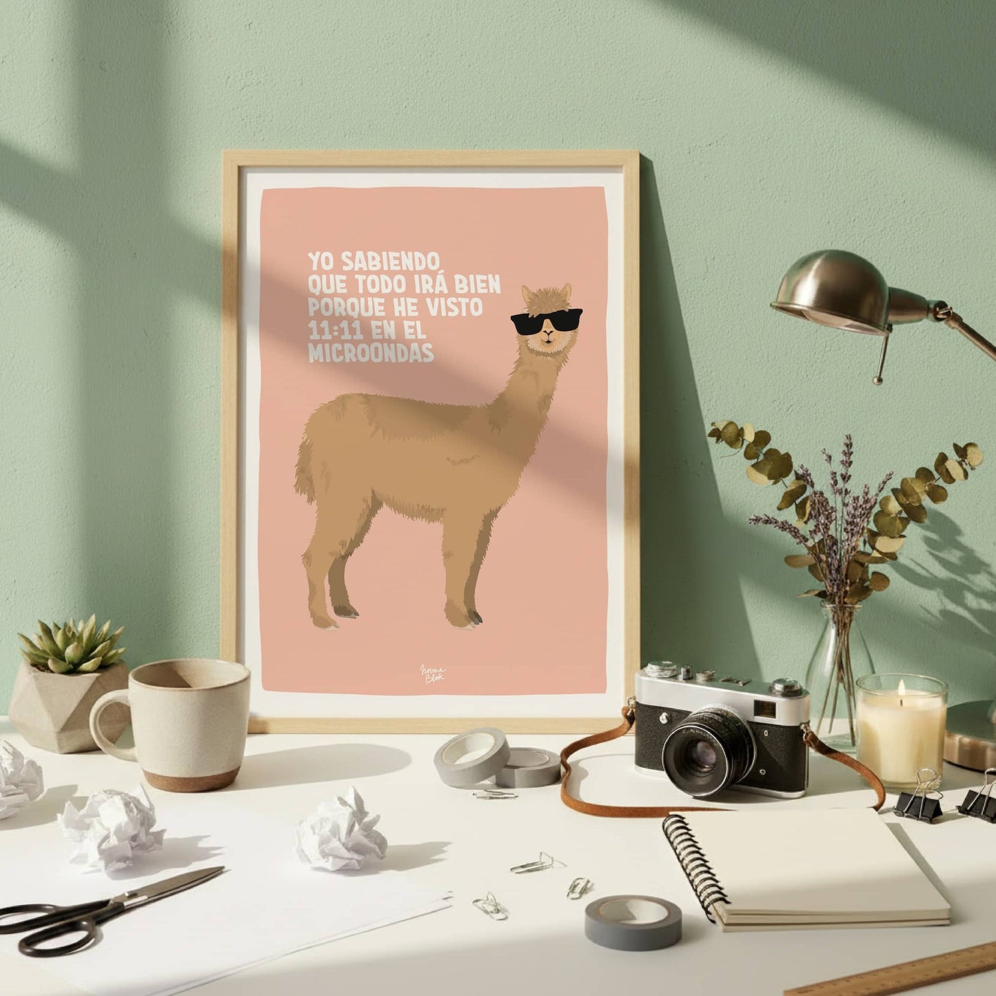 La lámina de la alpaca en un marco de madera natural apoyada en una pared verde oscura. Está sobre un escritorio desordenado con libretas y bolis, mostrando cómo este diseño de afirmación caótica encaja perfectamente en un espacio de trabajo real, ocupado y del día a día.