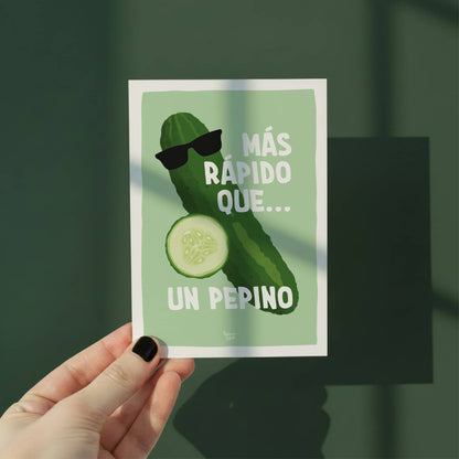 Una mano sosteniendo la postal del pepino frente a un fondo verde para mostrar su tamaño real y cómo los tonos verdes vivos destacan como un pequeño oasis de color por normablok.