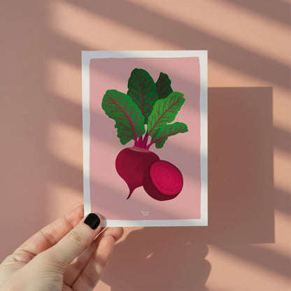 Una mano sosteniendo la mini lámina de la remolacha para mostrar el tamaño A6 y el fondo rosado vibrante, ideal para usar como ficha de receta o decoración.