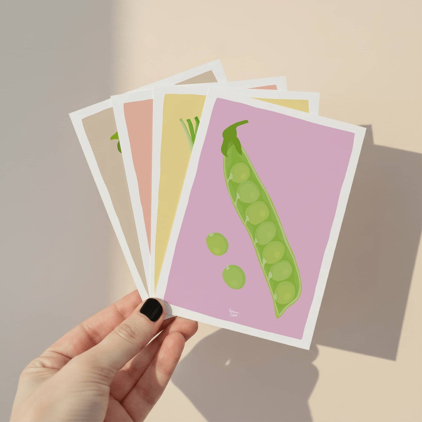 A close-up of a hand holding the full set of four vegetable mini prints to show the high-quality paper, the A6 size, and the variety of colorful backgrounds.