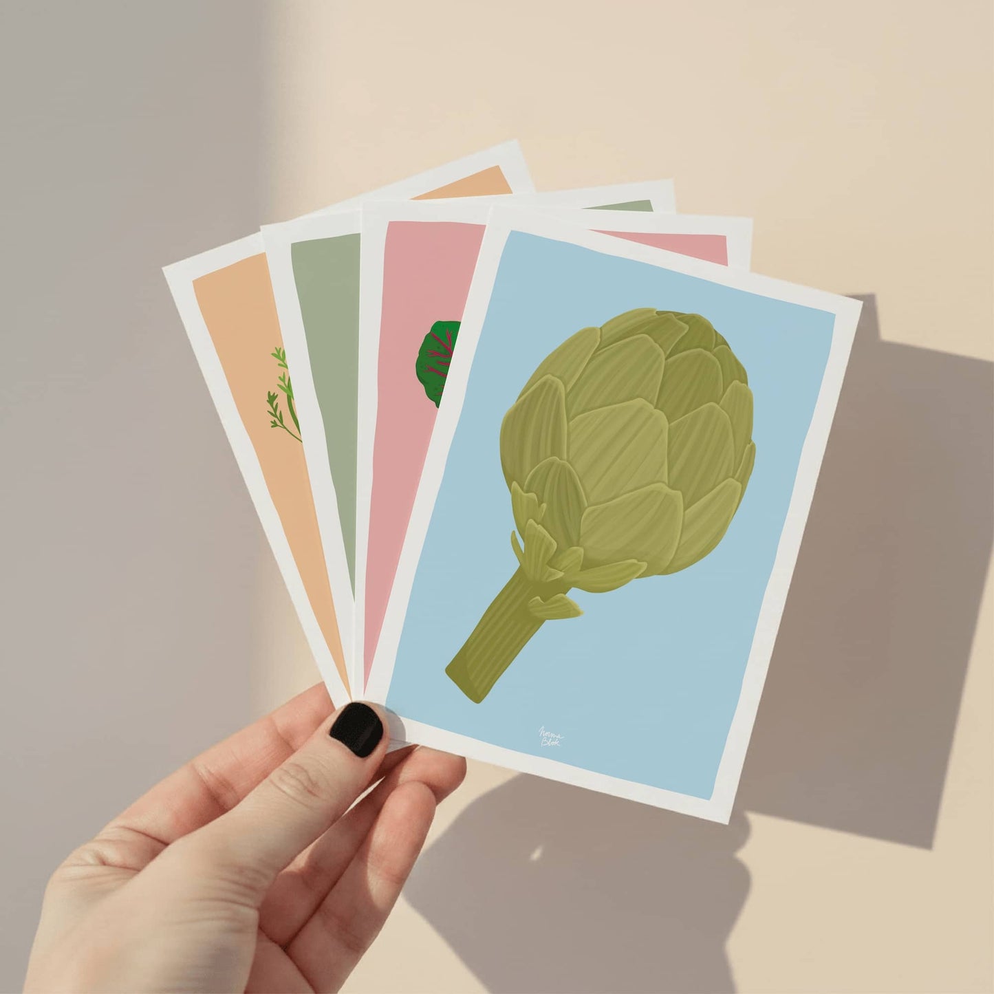 Una mano sosteniendo el pack completo de las cuatro mini láminas de verduras, mostrando la colección junta y el acabado de calidad de la ilustración digital.