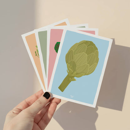 Una mano sosteniendo el pack completo de las cuatro mini láminas de verduras, mostrando la colección junta y el acabado de calidad de la ilustración digital.