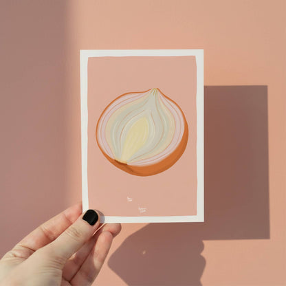 A hand holding the onion mini print with its soft pink background, highlighting the figurative digital style and the white perimeter border.