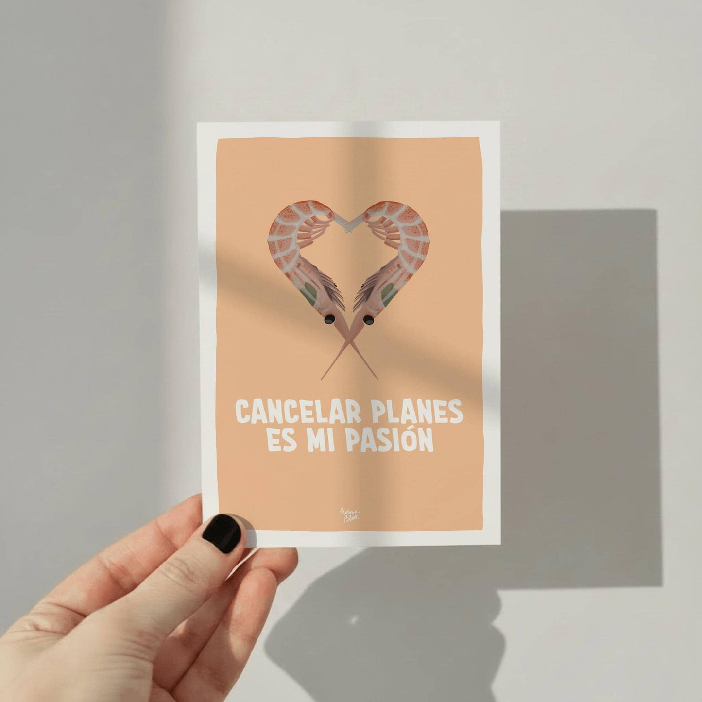 Fotografía de una mano sujetando por un borde la postal de Normablok ('cancelar planes es mi pasión') contra un fondo de estudio blanco y neutro. Se aprecia el tamaño real de la tarjeta, la textura del papel mate y la ilustración de las gambas con el fondo naranja.
