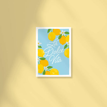 Postal A6 con fondo azul cielo que muestra el texto "la dolce vita" en letra ligada central rodeado de limones y flores de limonero en las esquinas por normablok.
