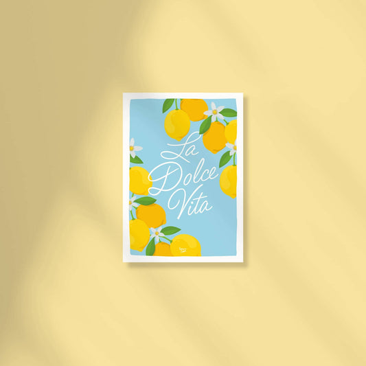 Postal A6 con fondo azul cielo que muestra el texto "la dolce vita" en letra ligada central rodeado de limones y flores de limonero en las esquinas por normablok.