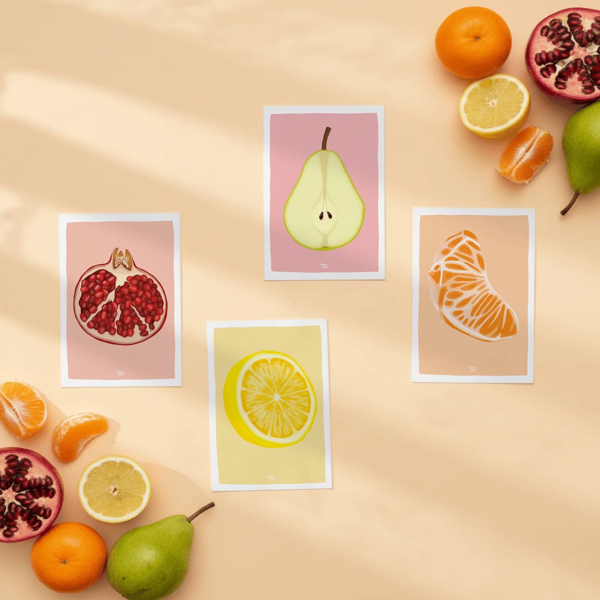 Las cuatro mini láminas de frutas (granada, limón, mandarina y pera) de Normablok dispuestas sobre un fondo beige y rodeadas de piezas de fruta real. Se aprecia el detalle de la ilustración digital, el tamaño A6 y el marco blanco perimetral diseñado para facilitar el enmarcado.