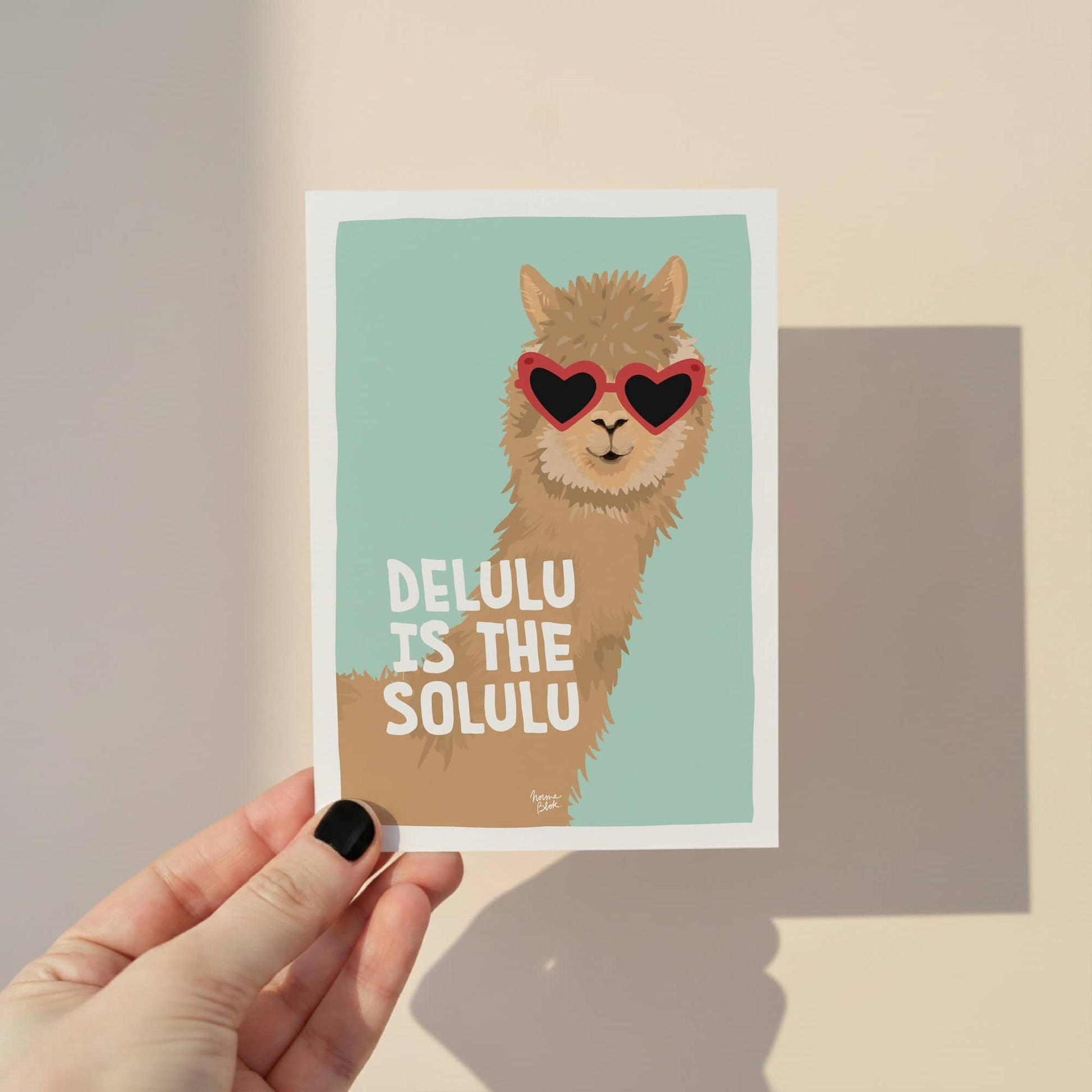 A6 postcard with the text 'Delulu is the solulu' featuring a medium shot of an alpaca wearing heart-shaped sunglasses against a vibrant turquoise background by Normablok.