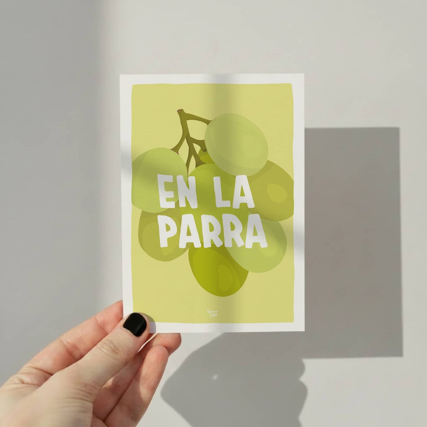 A hand holding the A6 postcard against a wooden desk to show its size, highlighting the colorful and figurative art style of the grapes by normablok.