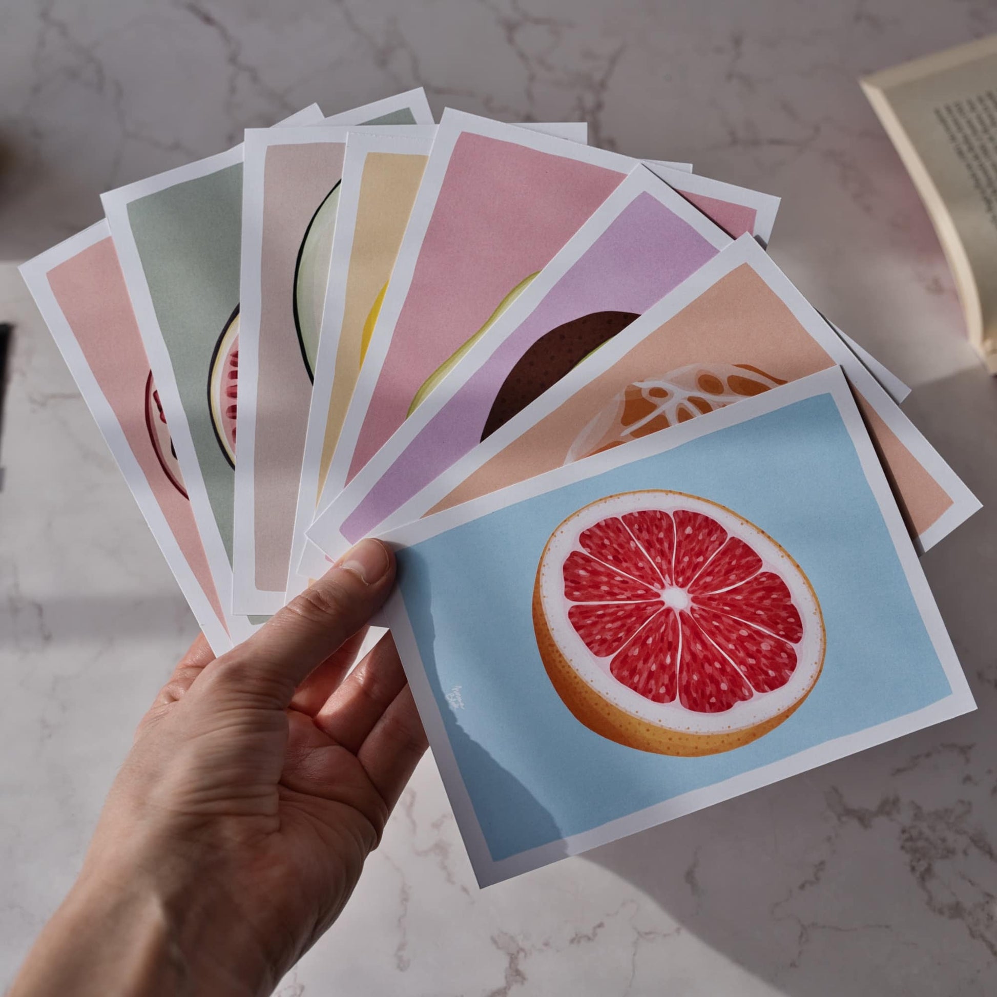 A close-up shot of a person's hand holding all eight fruit reading log cards spread out in a fan shape to showcase the entire collection. The vibrant digital illustrations of the pomegranate, fig, and other fruits are clearly visible against a soft background, showing the A6 size and the colorful, figurative art style. Each card is designed to help you keep track of your library and fight off that "book amnesia" we all know too well.