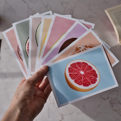 A close-up shot of a person's hand holding all eight fruit reading log cards spread out in a fan shape to showcase the entire collection. The vibrant digital illustrations of the pomegranate, fig, and other fruits are clearly visible against a soft background, showing the A6 size and the colorful, figurative art style. Each card is designed to help you keep track of your library and fight off that "book amnesia" we all know too well.