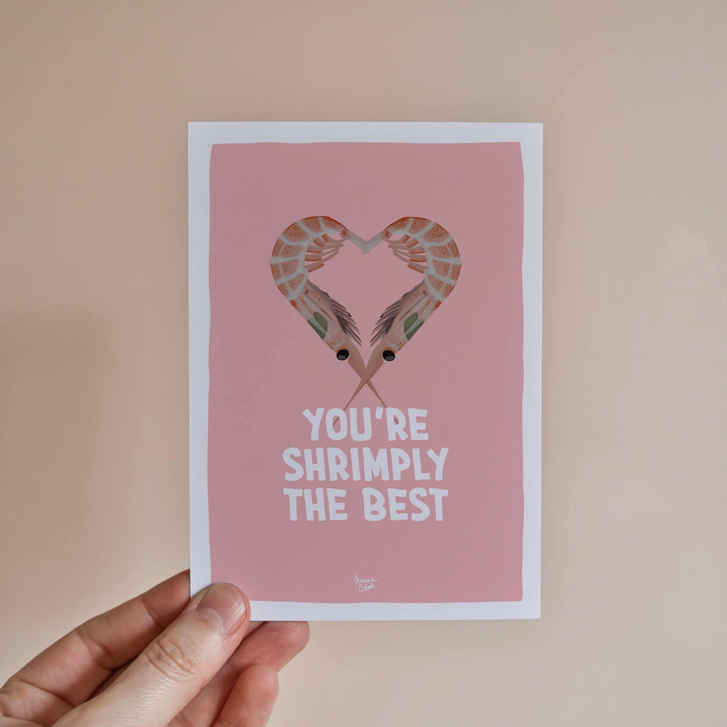 A hand holding the A6 shrimp pun card to showcase its size and the vibrant contrast between the orange shrimps and the soft pink background by normablok.