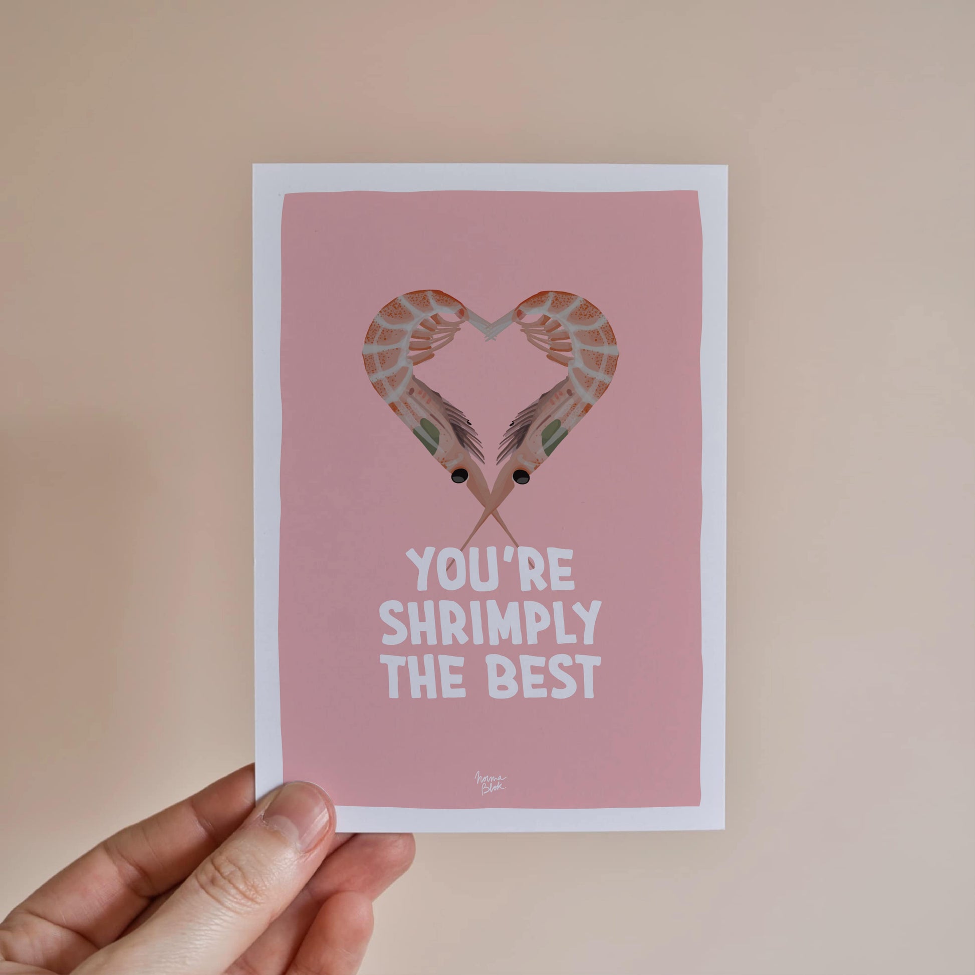 A hand holding the A6 shrimp pun card to showcase its size and the vibrant contrast between the orange shrimps and the soft pink background by normablok.