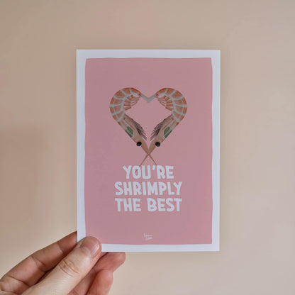 A hand holding the A6 shrimp pun card to showcase its size and the vibrant contrast between the orange shrimps and the soft pink background by normablok.