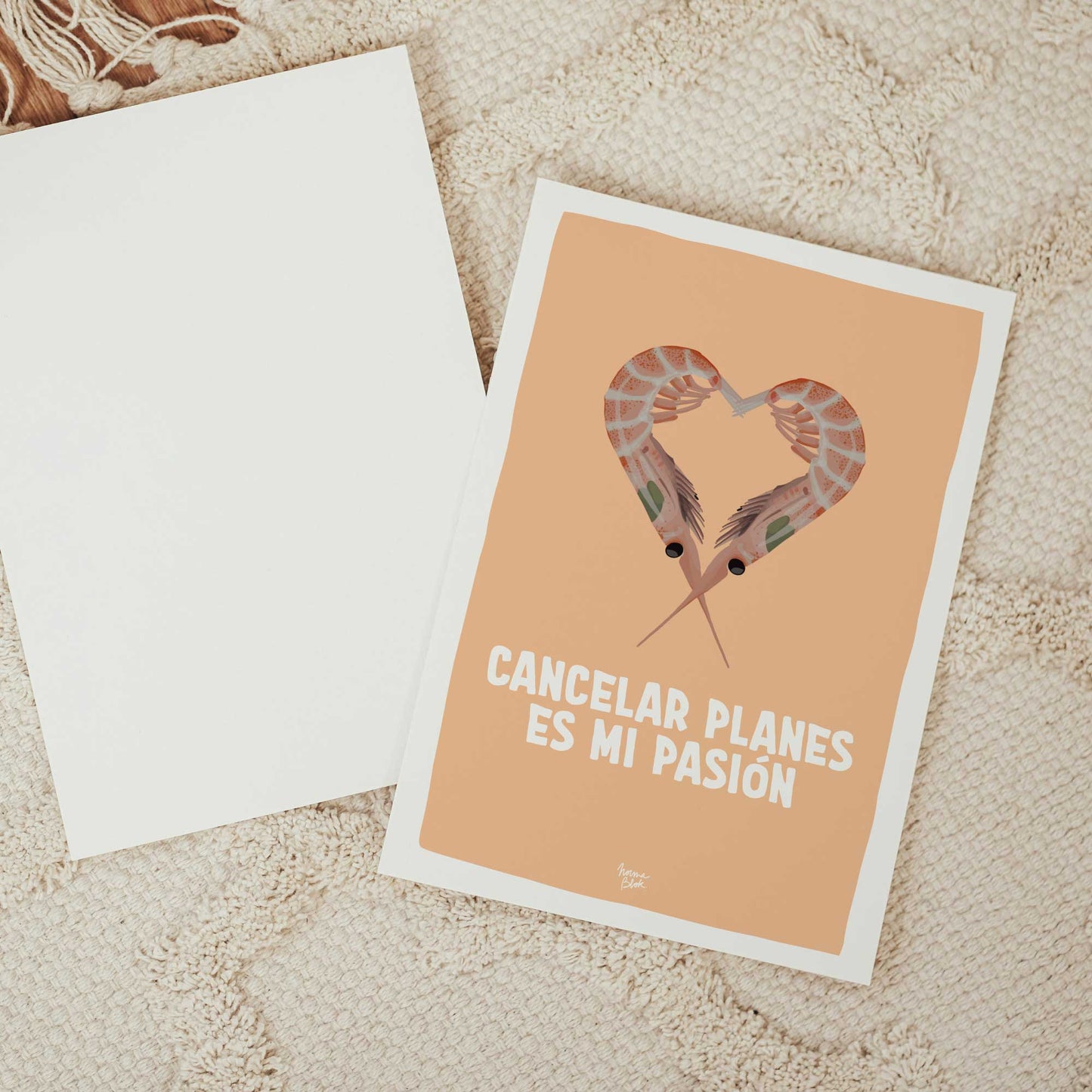Composición 'flat lay' o vista cenital sobre una alfombra de textura beige. A la izquierda, la lámina de Normablok 'cancelar planes es mi pasión' con su diseño de gambas y fondo naranja. A la derecha, otra postal idéntica dada la vuelta, mostrando el reverso de papel blanco en blanco