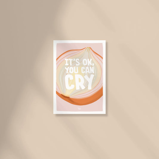 A soft pale pink A6 postcard featuring a digital illustration of a white onion sliced in half to show its internal layers, with the text "it's ok you can cry" written in a modern, casual font by normablok.