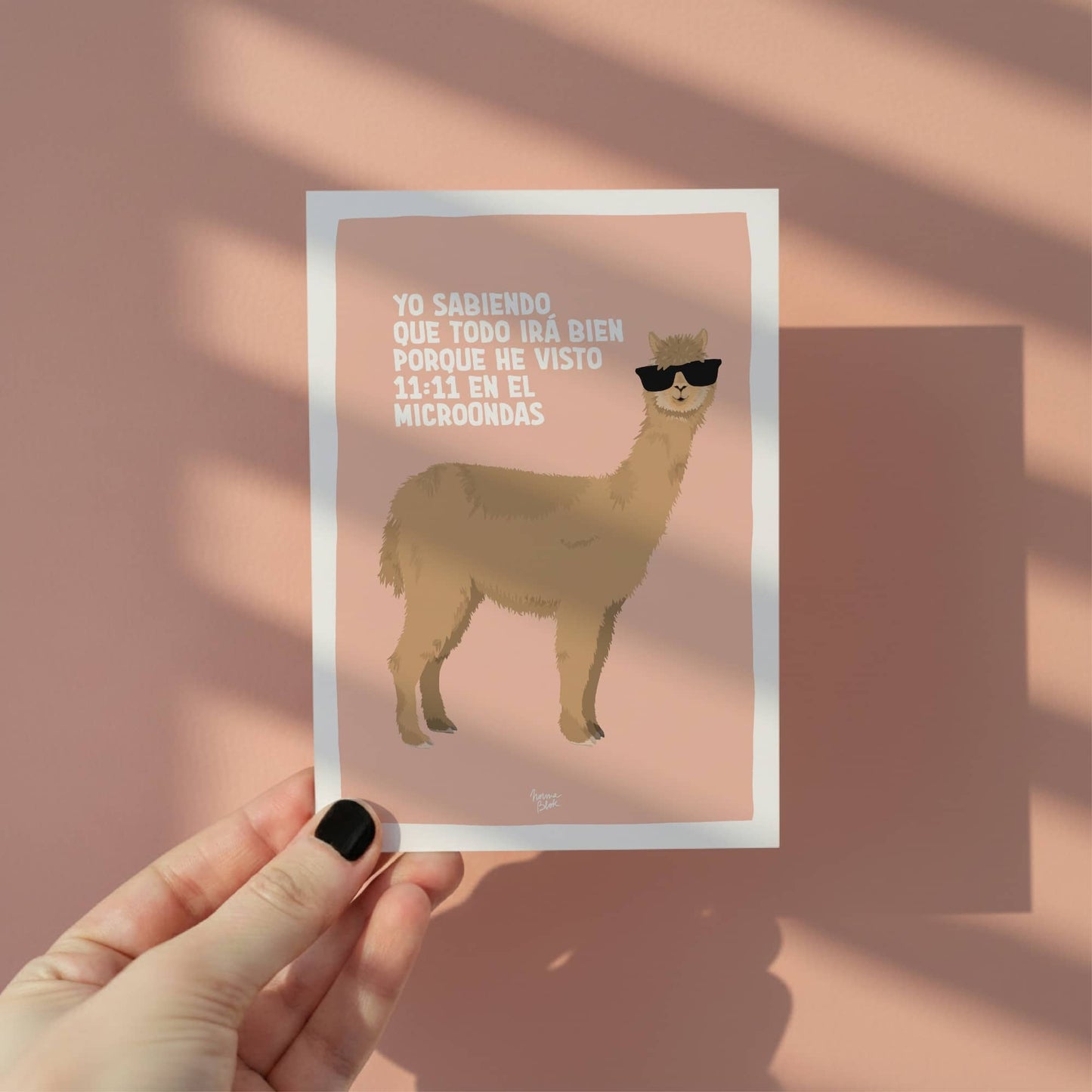 Primer plano de una mano sosteniendo la ilustración de la alpaca con gafas de sol, permitiendo observar la calidad de la impresión, el tamaño postal A6 y el marco blanco que facilita el enmarcado del diseño figurativo.