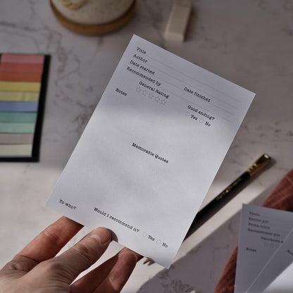 A close-up of a hand holding the English version of the fruit-themed reading log card. The photo highlights the "Book Review" section where you can track ratings and memorable quotes to prevent "book amnesia". This A6 bookmark features a vibrant, figurative fruit illustration on the front, making it a stylish and functional literary gift. By normablok.