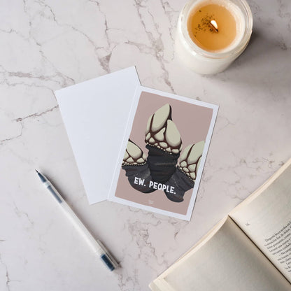 Top-down view of an "Ew. People." barnacle postcard on a white marble surface next to a lit candle, a sleek pen, and the clean blank back of the postcard by Normablok