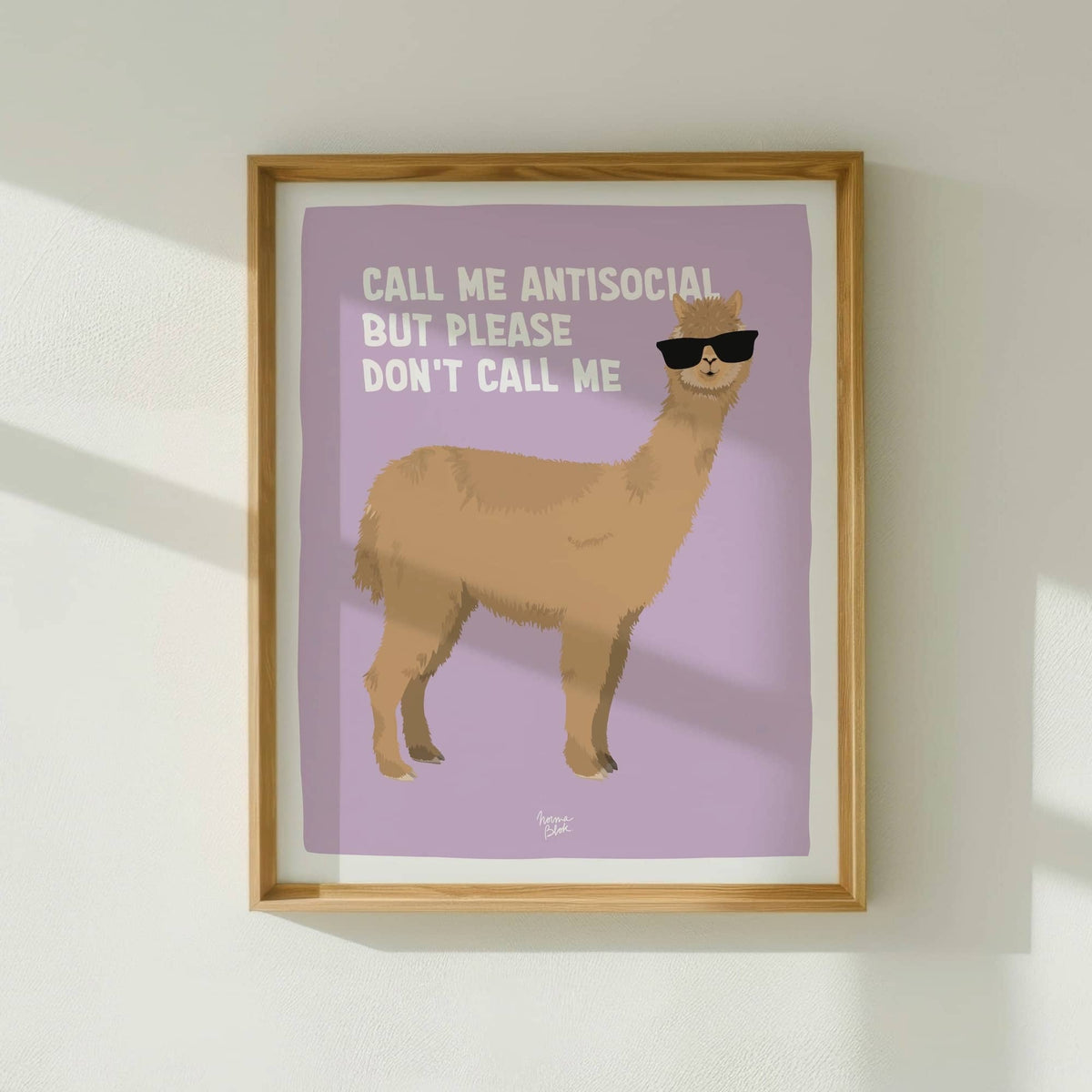 The antisocial alpaca art print framed in light wood, hanging on a white wall with natural window light by Normablok.