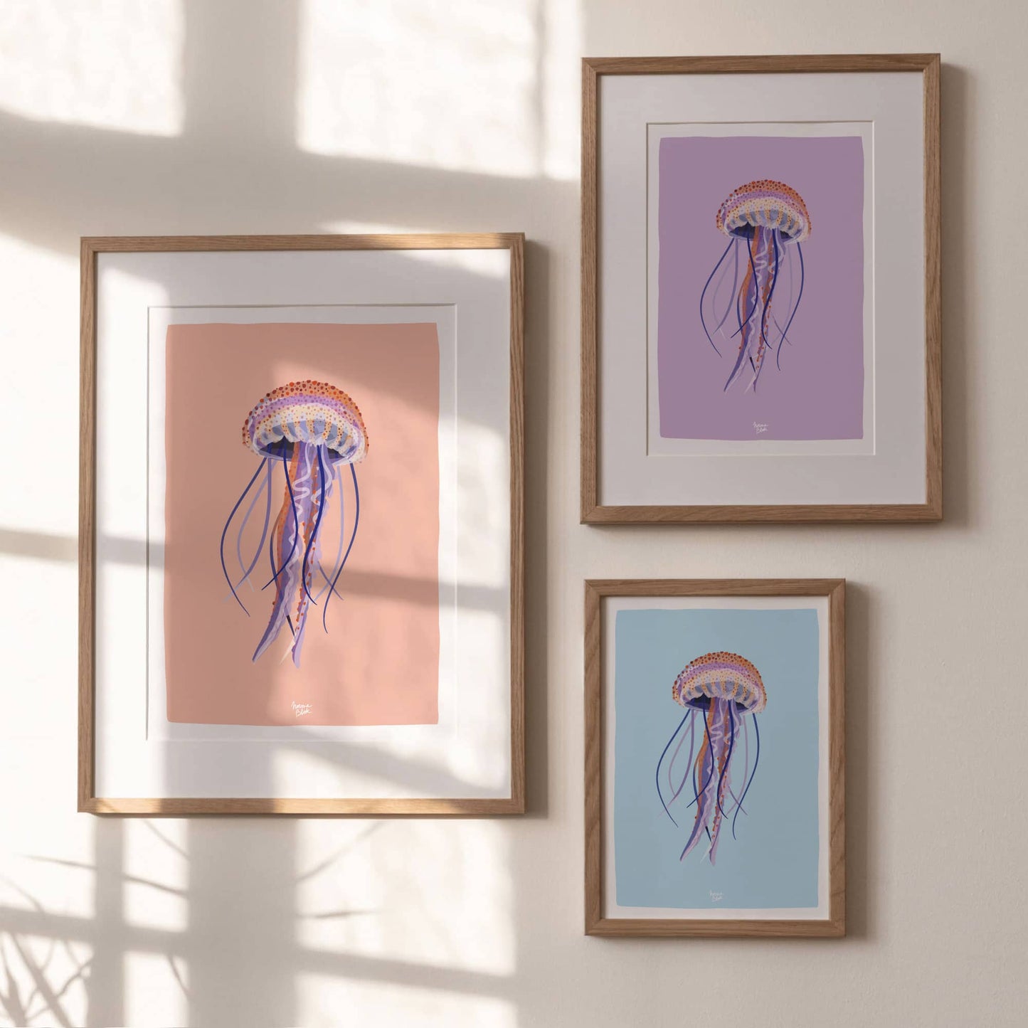 A gallery wall setup featuring three framed jellyfish prints with different backgrounds, orange, lilac, and blue, hung neatly on a plain white wall. This display shows how well the collection works together to bring a pop of color to any room.