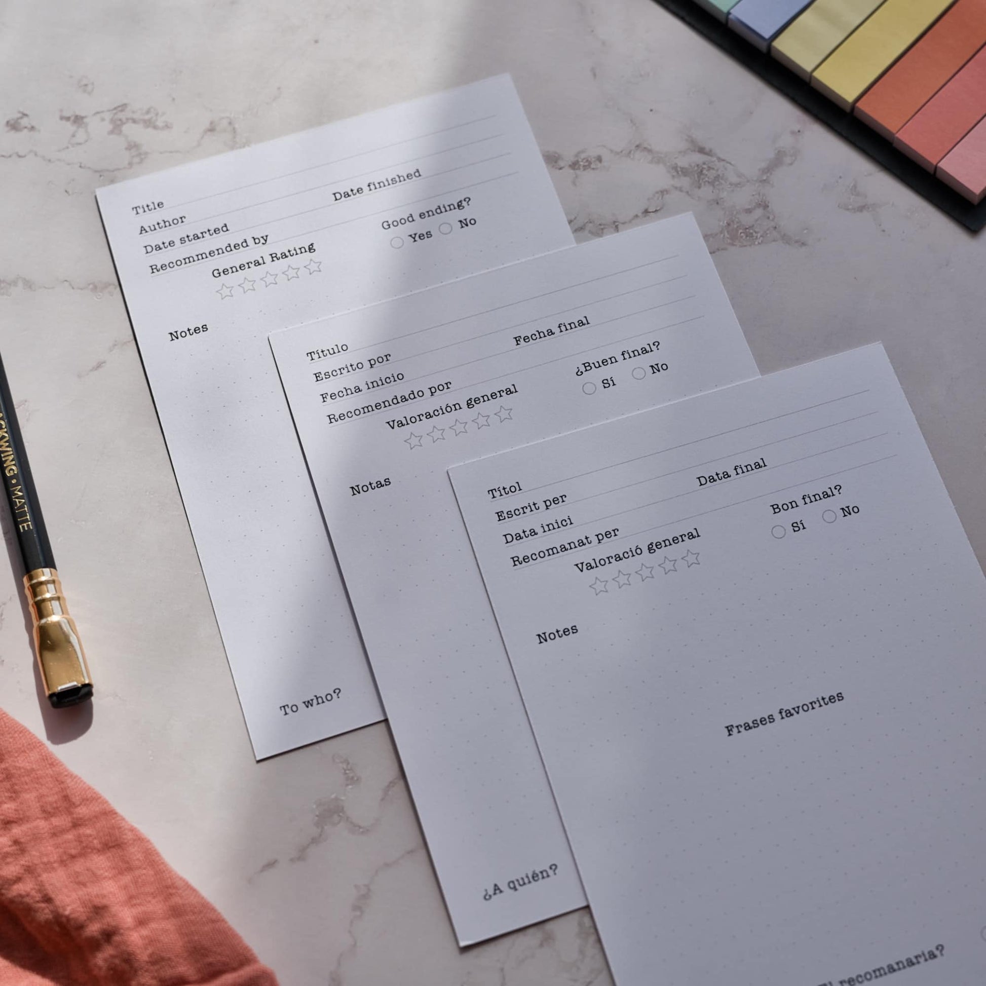 A top-down view of three fruit reading log cards laid out on a table, showcasing the multilingual options in Spanish, Catalan, and English. The vibrant digital fruit illustrations are visible alongside the functional back template for book tracking. It’s a great way to see how these A6 bookmarks help you organize your library in your preferred language. By normablok.