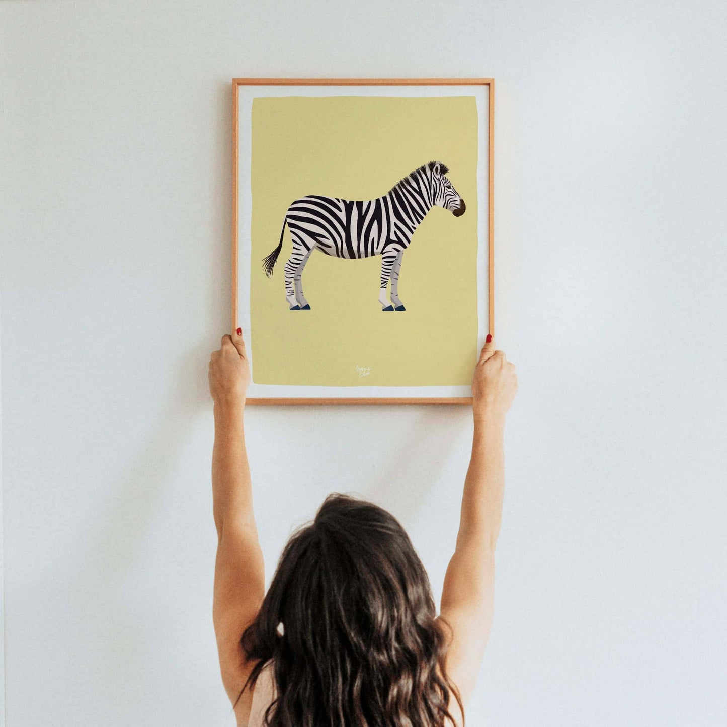A woman's hands hanging an A3-sized framed zebra art print on a wall. This real-life reference helps customers understand the physical scale and impressive size of the A3 print, making it easier to visualize its presence in a bedroom or playroom.