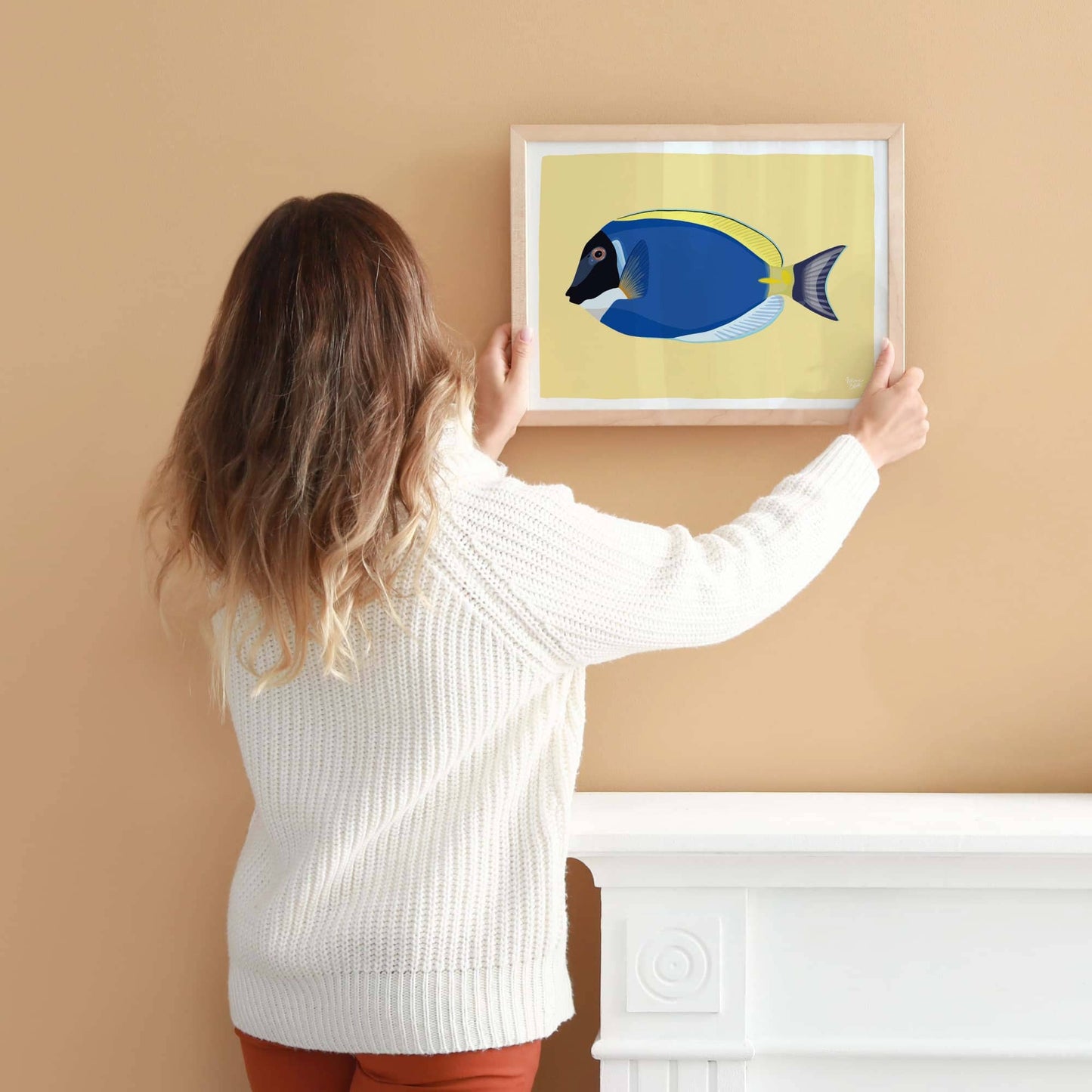 A woman hanging an A4-sized framed Blue Tang fish illustration on a light coral-colored wall. Shows the scale for wall decoration.