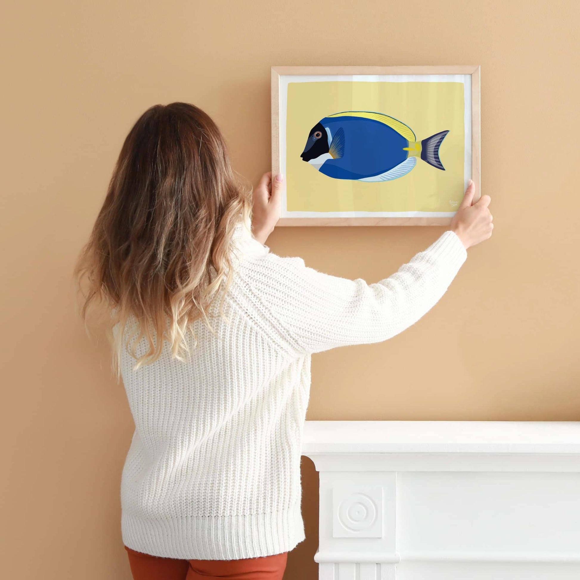 A woman hanging an A4-sized framed Blue Tang fish illustration on a light coral-colored wall. Shows the scale for wall decoration.