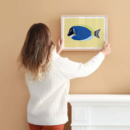 A woman hanging an A4-sized framed Blue Tang fish illustration on a light coral-colored wall. Shows the scale for wall decoration.