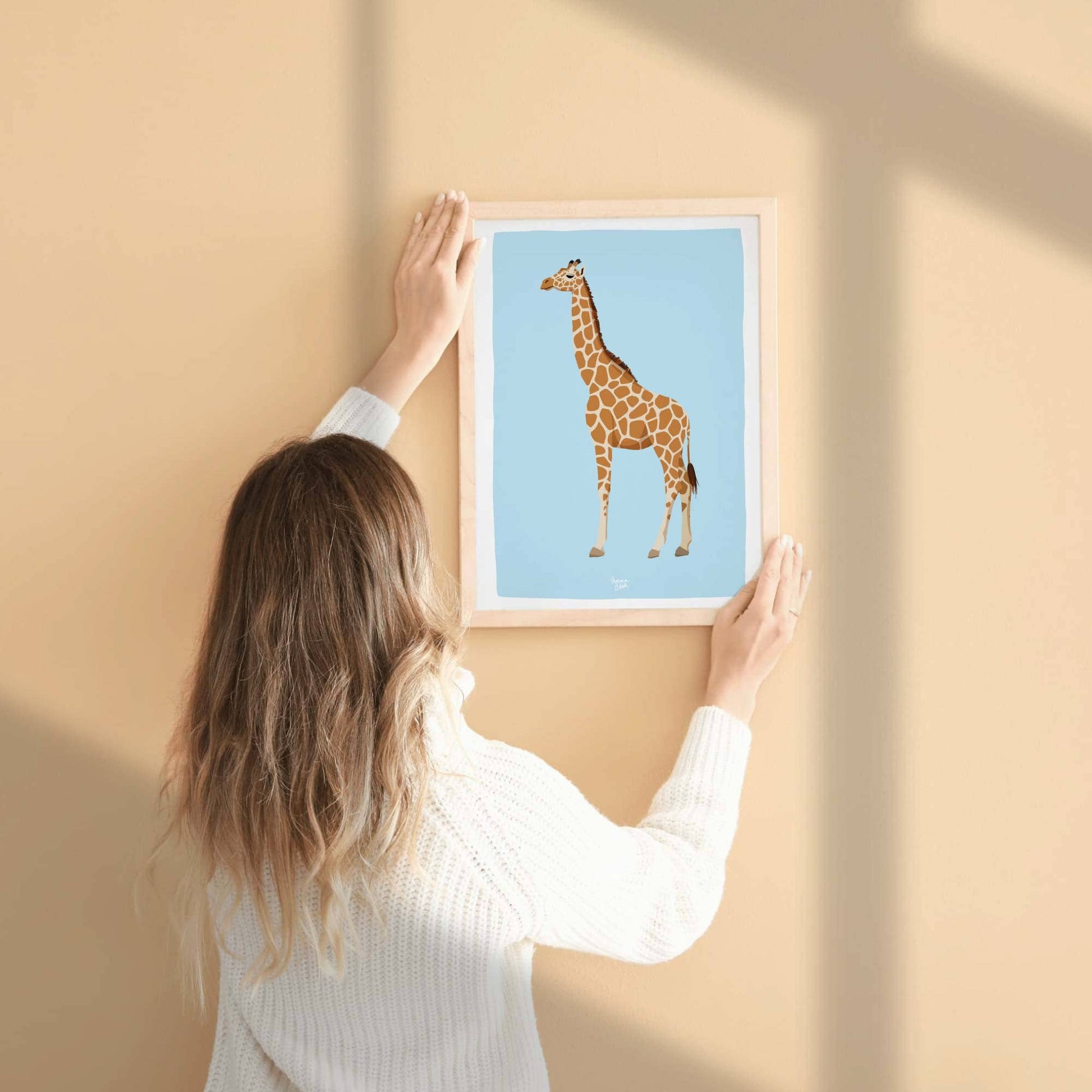 A woman hanging an A4-sized framed giraffe print on a warm, salmon-colored wall. This real-life perspective illustrates the perfect scale of the wall art and how vibrant home decor colors harmonize beautifully with modern, hand-drawn animal illustrations.