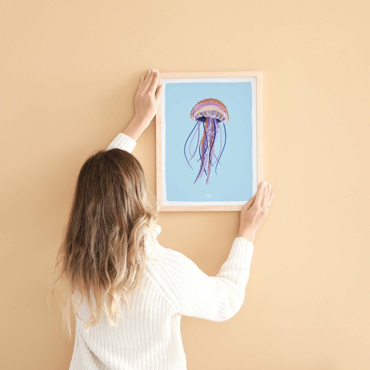 A woman in the process of hanging an A4-sized framed jellyfish print on a wall. This image helps you get a real sense of the print's scale and how it sits comfortably in a modern, lived-in home environment.