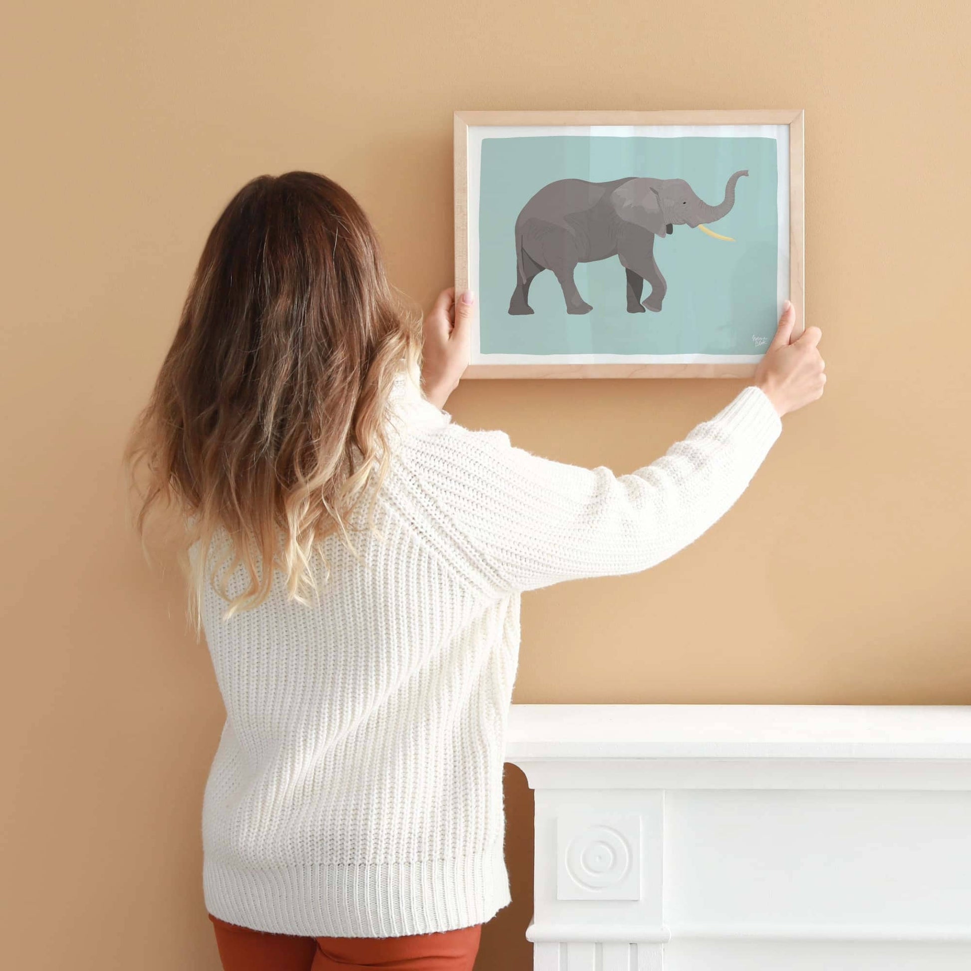 Una mujer cuelga la lámina del elefante enmarcada en tamaño A4 en una pared neutra. Sus manos sirven como referencia real de la escala física, el tamaño y las proporciones de la ilustración impresa, ayudando a visualizar cómo queda en una composición de pared actual.